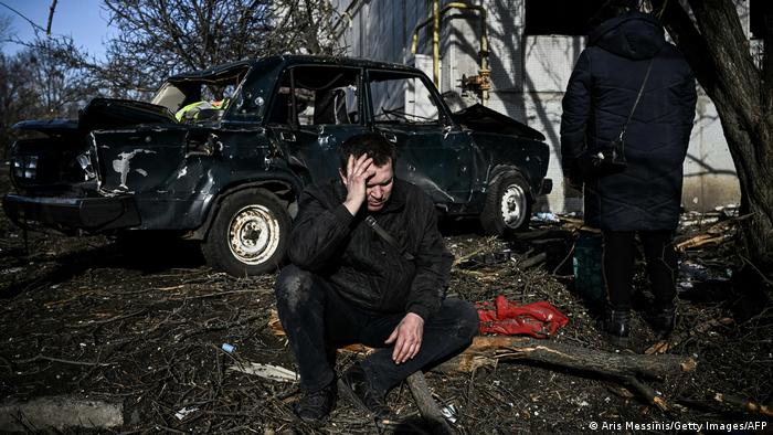 Мужчина сидит рядом с разрушенным автомобилем и жилым зданием после ракетного удара и атак боевых вертолетов в городе Чугуев в Харьковской области